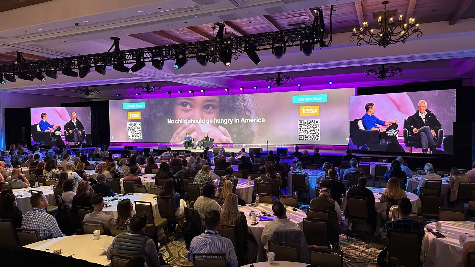Corporate panel on stage with live camera feeds on side LED screens above a seated hotel ballroom audience